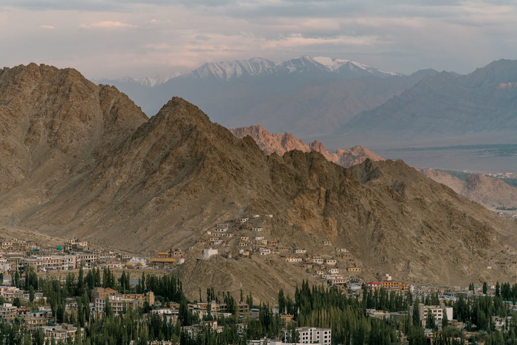 Drone view of town located amidst green trees near rocky mountains with buildings located in highlands against snowy peaks in foggy weather