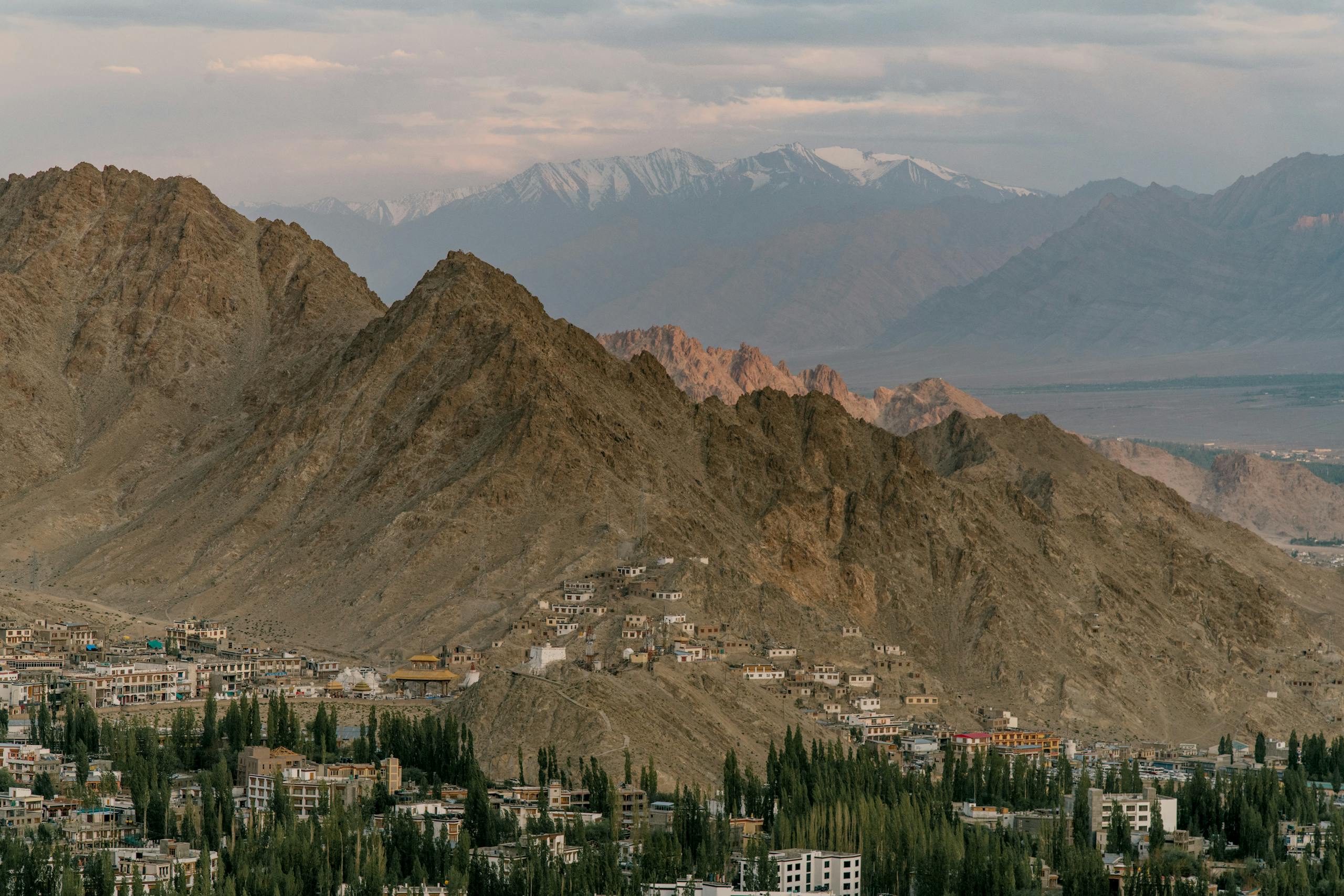 Drone view of town located amidst green trees near rocky mountains with buildings located in highlands against snowy peaks in foggy weather